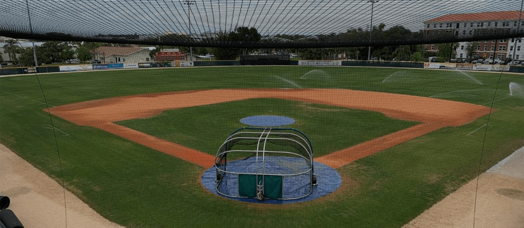 Panoramic view from behind home plate showing protective baseball field netting, a batting cage over home plate, and the outfield